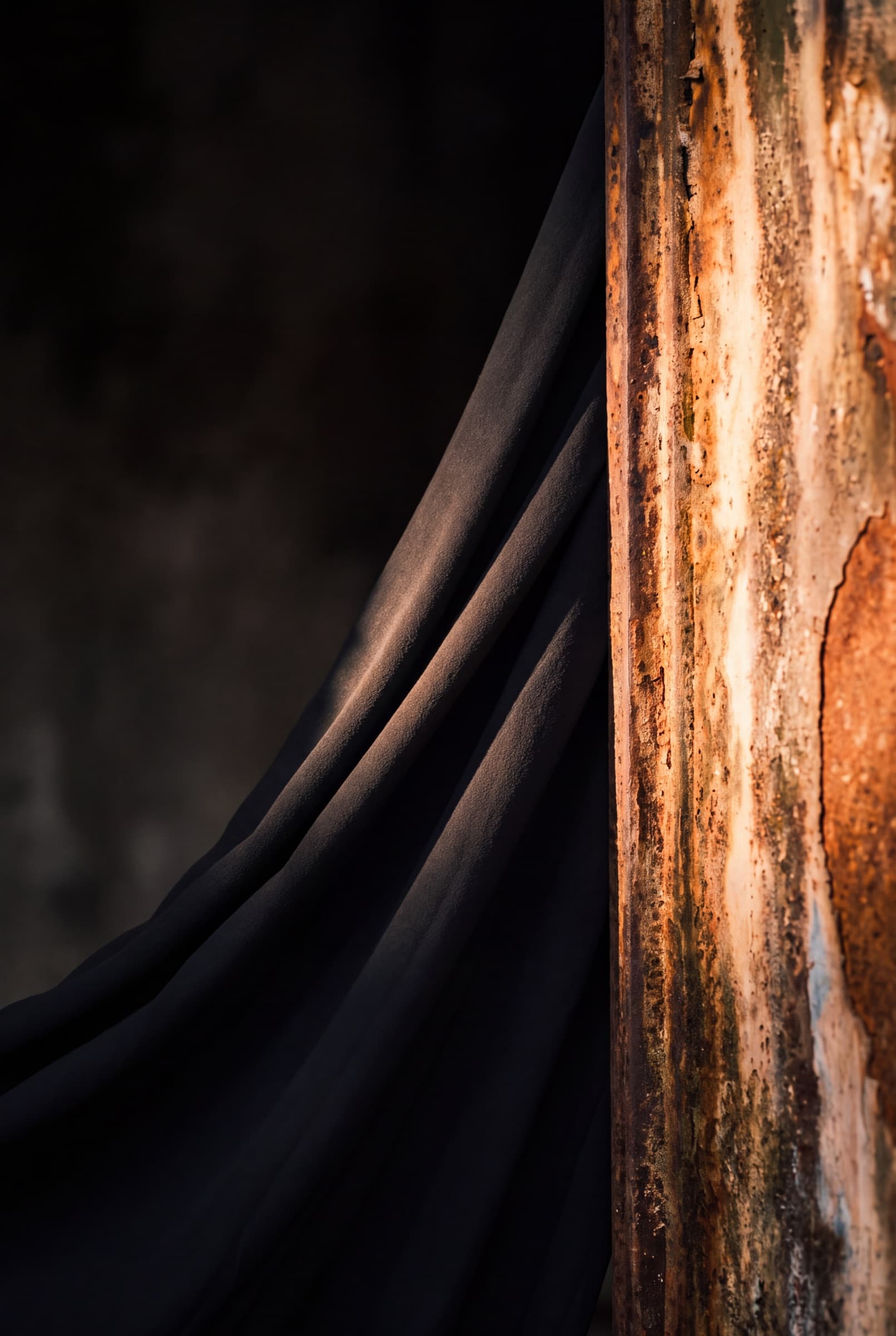 Black crepe fabric sliding off an oxidised iron plate, a line of light along one fold.